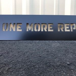 May include: Black metal sign with the words "ONE MORE REP" cut out in a stencil style.