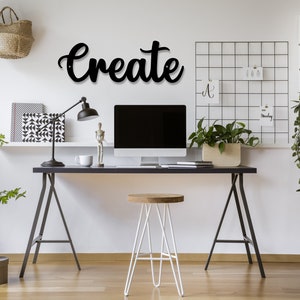 May include: A black metal word art sign that says "Create" is hanging on a white wall above a black desk with a white computer monitor, a white stool, and a black lamp. There is a white shelf with books and a white ladder shelf with plants and a camera on it.