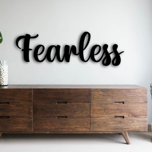 Puede incluir: Arte de pared de madera negra con la palabra "Fearless" escrita en cursiva. El letrero está montado en una pared blanca sobre un mueble de madera de estilo moderno. Una gran hoja verde está en un jarrón transparente.