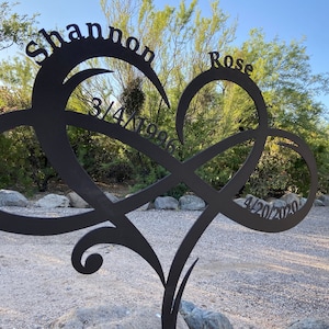 May include: A black metal heart-shaped sculpture with the names "Shannon" and "Rose" and the dates "3/4/1986" and "4/20/2020" inscribed on it.