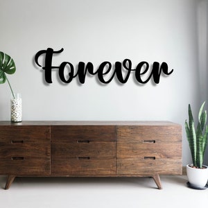 May include: A wooden dresser with six drawers, a black metal word art sign that says "Forever" on a white wall, a potted green plant, and a vase with white stones on a white surface.