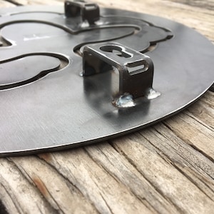 Puede incluir: Una placa de metal con recortes en forma de flor de lis. La placa tiene cuatro soportes metálicos unidos a la parte posterior.