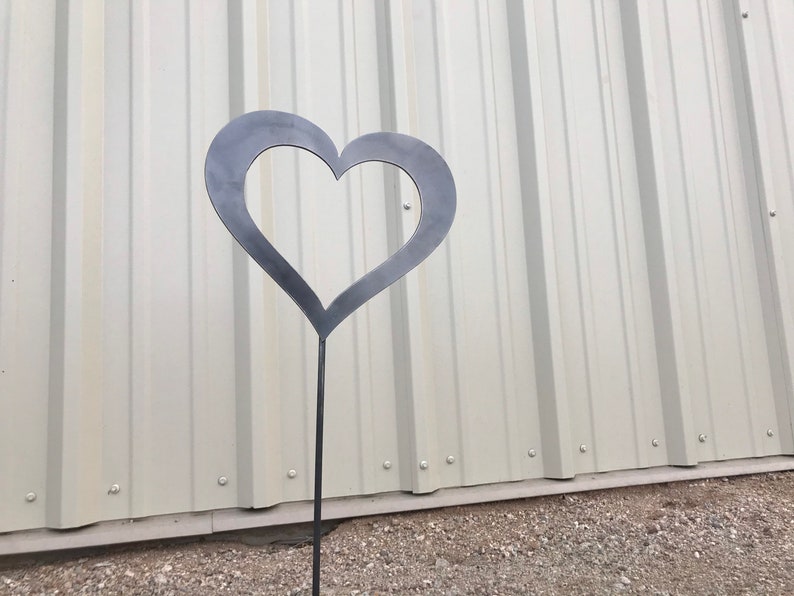 Pu&ograve; includere: Un paletto da giardino in metallo nero a forma di cuore con un'estremit&agrave; appuntita. Il paletto &egrave; davanti a un muro di metallo bianco.