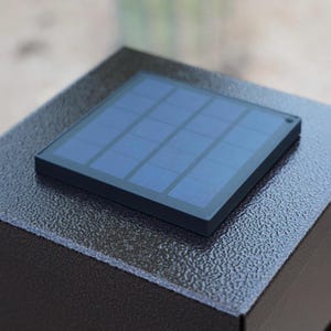 May include: A close-up of a black solar panel with a grid of blue cells. The panel is square and set on a textured, dark gray surface. The solar panel is a component of an outdoor light fixture.