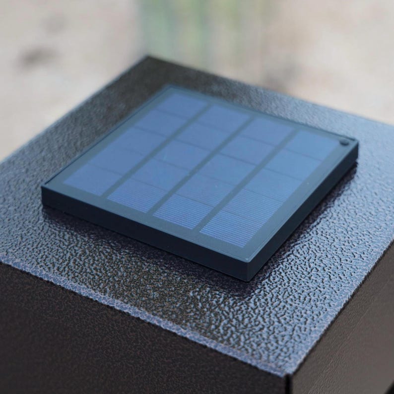 May include: A close-up of a black solar panel with a grid of blue cells. The panel is square and set on a textured, dark gray surface. The solar panel is a component of an outdoor light fixture.