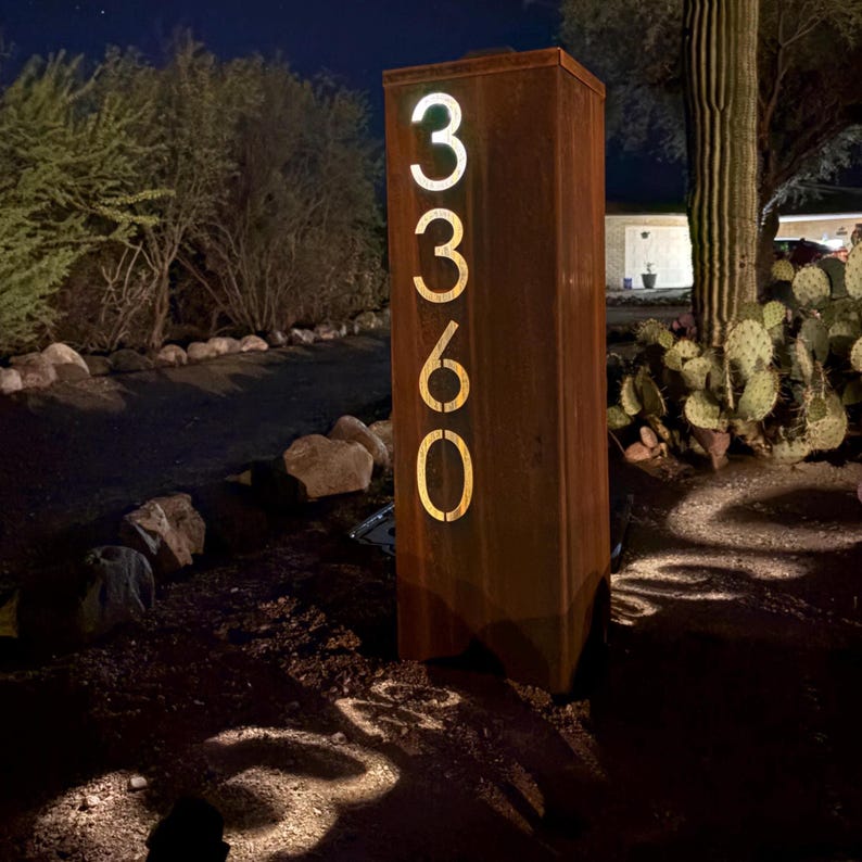 May include: A rectangular, weathered metal address sign with the numbers 3360 illuminated in a warm, inviting glow. The sign is set in a desert landscape at night, with a cactus and other desert plants in the background.