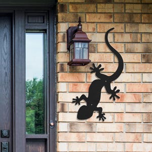 Puede incluir: Decoración de pared de metal negro con forma de gecko, fijada a una pared de ladrillos. El gecko tiene una cola larga y curvada y pies detallados. Una linterna exterior marrón está montada encima del gecko. La pared de ladrillo es de ladrillos marrones claros y oscuros.