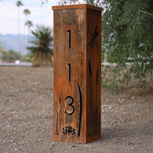 Könnte beinhalten: Ein rechteckiger, verwitterter Adresspfosten aus Metall mit der ausgeschnittenen Nummer "113". Der Pfosten zeigt ein Libellen-, Schilf- und Lotusblumen-Design. Der Pfosten hat eine rustikale, orangebraune Farbe.