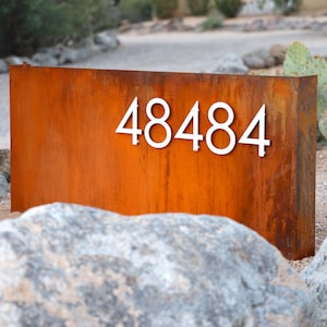 May include: A rectangular, weathered metal mailbox with the number "48484" in white, mounted on the front. The mailbox has a rusty orange color and is set in a desert landscape with rocks and cacti.