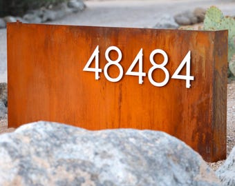 May include: A rectangular, weathered metal mailbox with the number "48484" in white, mounted on the front. The mailbox has a rusty orange color and is set in a desert landscape with rocks and cacti.