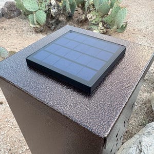 May include: A solar-powered outdoor light fixture with a textured, dark brown finish. The fixture has a rectangular shape with a solar panel on top. The panel is black with blue cells. The background shows desert plants.