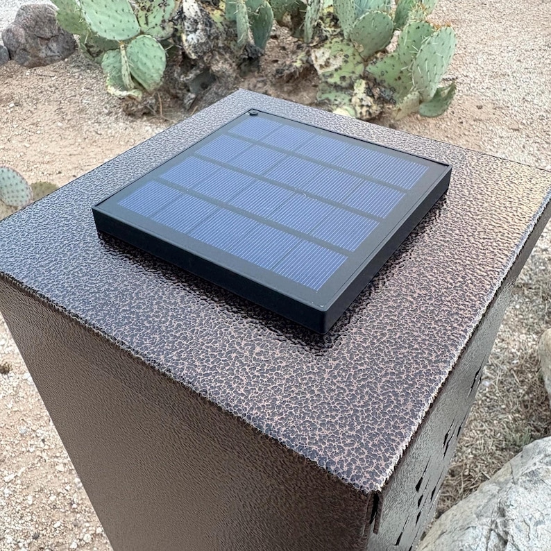 May include: A solar-powered outdoor light fixture with a textured, dark brown finish. The fixture has a rectangular shape with a solar panel on top. The panel is black with blue cells. The background shows desert plants.