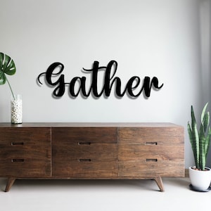 May include: Black wooden sign with the word "gather" in a cursive font, hanging on a white wall above a dark brown wooden dresser with six drawers.
