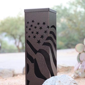 May include: A bronze-colored metal post with a cut-out American flag design. The flag features stars and stripes, with the stars at the top. The post is rectangular with a flat top and is set outdoors.