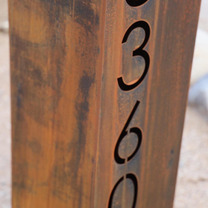 May include: A weathered, rectangular metal sign post with the numbers 360 cut out. The post has a rusty brown color with darker brown and black variations. The numbers are in a simple, sans-serif font.