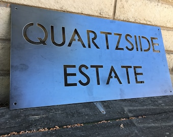 May include: A blue metal sign with the words "Quartzside Estate" cut out in black.