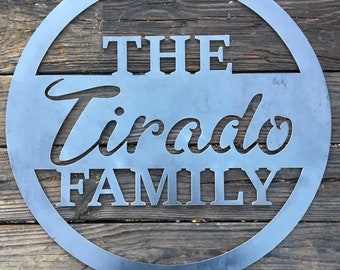 May include: A silver metal circle sign with the text "THE Tirado FAMILY" cut out. The sign is hanging on a wooden wall.