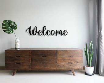 Peut inclure: Une pancarte en métal noir avec l'inscription « Bienvenue » en écriture cursive est accrochée à un mur blanc au-dessus d'une commode en bois brun foncé à six tiroirs. Une plante verte est dans un pot blanc sur le côté droit de la commode.