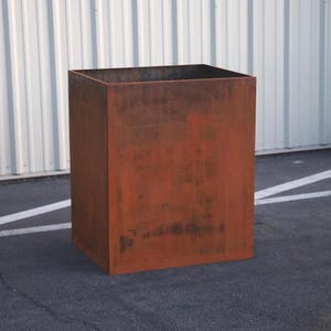 May include: A large, rectangular planter made of rusted metal. The planter has a square shape and is sitting on a gray asphalt surface.