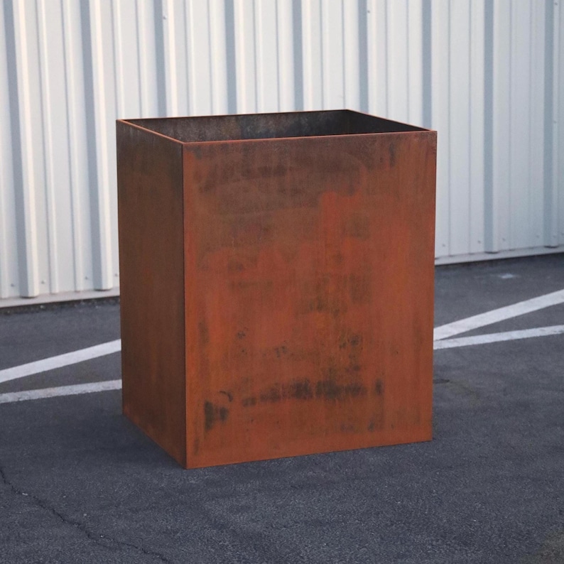 May include: A large, rectangular planter made of rusted metal. The planter has a square shape and is sitting on a gray asphalt surface.