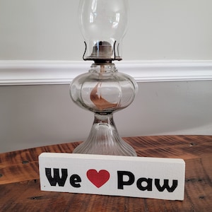 May include: A clear glass oil lamp with a bulbous base and a tall chimney. The lamp sits on a wooden surface next to a white wooden sign that reads "We ❤️ Paw" in black letters with a red heart.