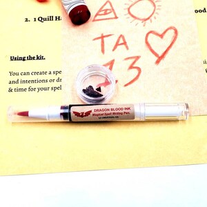 May include: A close-up of a kit for creating spells. The kit includes a quill, a bottle of red ink, a small jar of black powder, and a pen labeled "Dragon Blood Ink Magical Spell Writing Pen." The kit also includes a piece of paper with a triangle, a sun, a heart, and the letters "TA" and the number "13" drawn on it. The text "Using the kit. You can create a spell and intentions or draw & time for your spell" is written on the paper. The text "2. 1 Quill H:" is written on the paper.