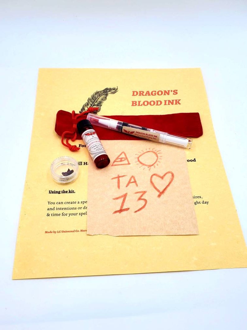 May include: A red velvet pouch containing a small bottle of red liquid labeled "Dragon's Blood Ink" and a white marker with the same label. A small piece of black resin is in a clear container. A piece of paper with the words "TA 13" written in red ink is on a yellow background. The text "Dragon's Blood Ink" is written in black on the yellow background. The text "Using the kit. You can create a spe and intentions or dr & time for your spel Made by LC Universal Co. Nori" is written in black on the yellow background.