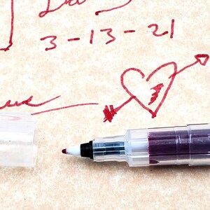 May include: A close-up of a clear, plastic pen with a burgundy ink cartridge. The pen is lying on a piece of paper with red ink markings, including the words "Party" and "3-13-21" and a heart with an arrow.