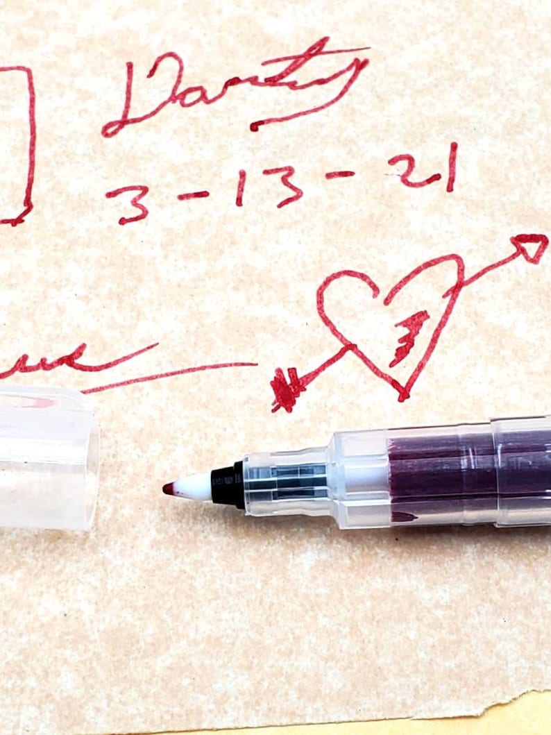 May include: A close-up of a clear, plastic pen with a burgundy ink cartridge. The pen is lying on a piece of paper with red ink markings, including the words "Party" and "3-13-21" and a heart with an arrow.