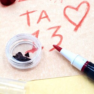 May include: A close-up of a small, clear container with a dark brown substance inside. A red paint marker is lying on a piece of paper with red drawings of a triangle, sun, heart, and the letters "TA 13".