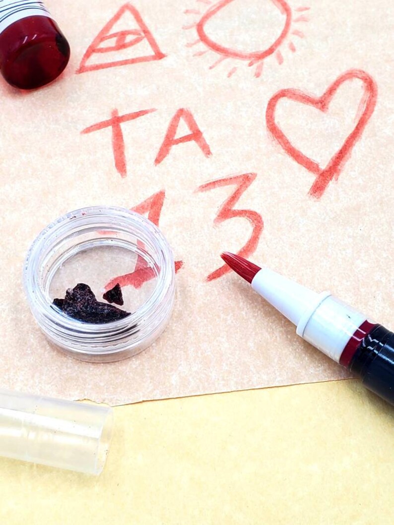 May include: A close-up of a small, clear container with a dark brown substance inside. A red paint marker is lying on a piece of paper with red drawings of a triangle, sun, heart, and the letters "TA 13".