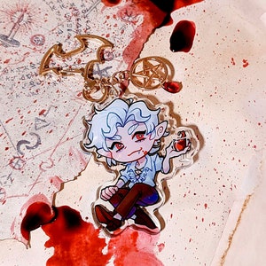 Könnte beinhalten: Ein weißer Vampircharakter mit roten Augen und blutigem Mund, der ein Glas mit roter Flüssigkeit hält. Der Charakter trägt ein weißes Hemd mit schwarzem Kragen und braune Hose. Der Charakter ist Teil eines Schlüsselanhängers mit einem goldenen Fledermaus- und Pentagramm-Anhänger.