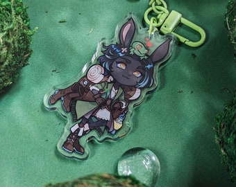 May include: A clear acrylic keychain featuring a cartoon character with bunny ears and a small frog. The keychain has a green clasp and a small frog-shaped charm. The background is green with moss and clear glass spheres.
