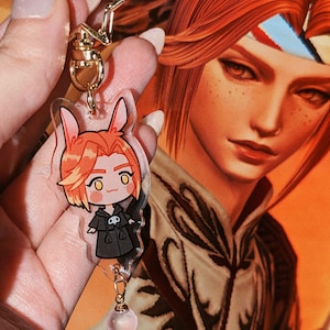 May include: A clear acrylic keychain with a cartoon character with orange hair and a black coat. The keychain has a gold star-shaped clasp and a small pink bead charm. The background is a portrait of a person with orange hair.