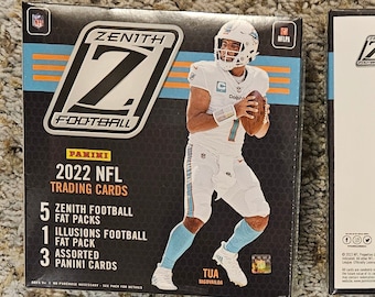 May include: A box of 2022 Panini Zenith Football trading cards featuring Tua Tagovailoa on the front. The box includes 5 Zenith Football Fat Packs, 1 Illusions Football Fat Pack, and 3 assorted Panini cards. The back of the box lists the card set details, including 100 base retail and 100 rookies retail.
