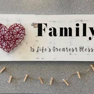 Puede incluir: Un letrero de madera blanco con las palabras "Family is Life's Greatest Blessing" en letras negras. Un corazón de cuerda roja está en el lado izquierdo del letrero. Una cuerda de mini pinzas para la ropa cuelga debajo del letrero.