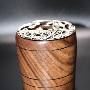 May include: A handcrafted wooden box with a decorative silver-colored lid. The box is cylindrical with a rich brown wood grain and black accent lines. The lid features an ornate, swirling design.