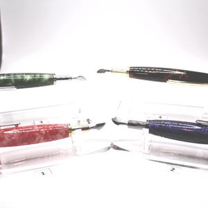 May include: Four fountain pens with unique designs are displayed on clear stands. The pens feature various colors, including green, red, brown, and blue, with gold and silver accents. Each pen has a distinct nib.