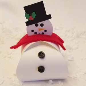May include: A white snowman-shaped gift box with a red felt scarf, black top hat, and green holly sprig. The snowman has black eyes, a carrot nose, and two black buttons.