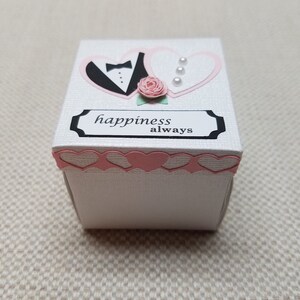 May include: White box with a pink and black heart design. The hearts are cut out and have a tuxedo and a dress on them. The box has the words "happiness always" on the front.