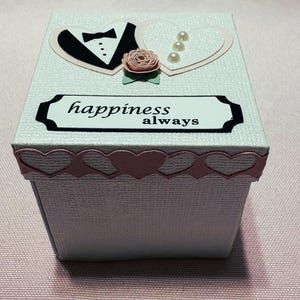 Pode incluir: Uma caixa de presente branca com uma superfície texturizada, decorada com um smoking preto e gravata borboleta, um coração, uma rosa rosa e as palavras "happiness always". A caixa tem uma borda de coração rosa.