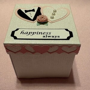 Może przedstawiać: Białe pudełko na prezent z różową obwódką w kształcie serca i pokrywką ozdobioną smokingiem, sercem i różą. Na pokrywce znajduje się również napis "happiness always" w czarnej ramce. Pudełko stoi na różowej powierzchni.