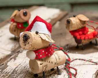 May include: Three cork dog ornaments with red and green accents. The dog on the left is wearing a white Santa hat. The dog in the center is wearing a red and white polka dot ribbon. The dog on the right is wearing a red pom-pom scarf.