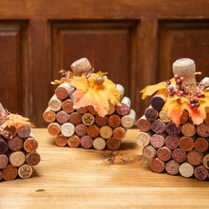 May include: Three handmade pumpkin decorations crafted from wine corks. Each pumpkin is a different size and features a cork stem, fall leaves, and decorative accents. The pumpkins are arranged on a wooden surface.