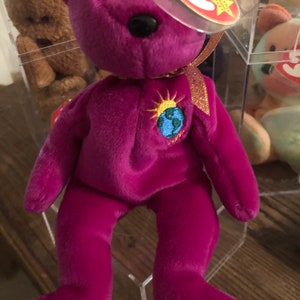 May include: A magenta plush bear with a black nose and embroidered globe and sun design on its chest. The bear has a red tag attached with a gold ribbon. Other plush toys are visible in the background.
