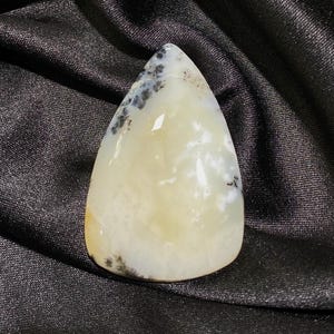 May include: A teardrop-shaped gemstone with a creamy white base and black speckles. The polished stone has a translucent quality, with a smooth surface. The gemstone is set against a black, silky background.