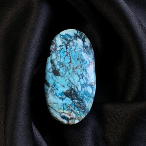 May include: An oval-shaped gemstone with a striking pattern of turquoise, black, and brown hues. The polished stone is set against a backdrop of black fabric, highlighting its natural colors and textures. The gemstone's unique design makes it a standout piece.