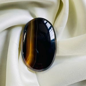 May include: An oval-shaped gemstone with a polished surface. The stone features a striking pattern of dark brown, amber, and black bands. The gemstone is set against a backdrop of white fabric.