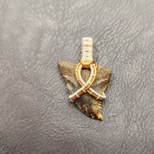 May include: A fossilised shark tooth pendant with a wire-wrapped design. The wire is wrapped in a criss-cross pattern and is made of silver and copper wire.