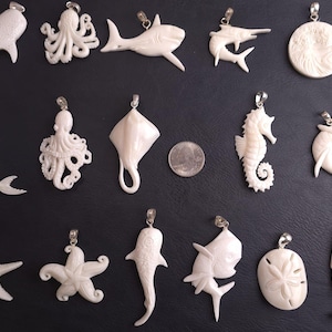 Puede incluir: Una colección de 15 colgantes tallados en hueso blanco con diferentes criaturas marinas, incluyendo una sirena, un pulpo, una raya, un caballito de mar, una estrella de mar y peces. Los colgantes están todos sobre un fondo negro.